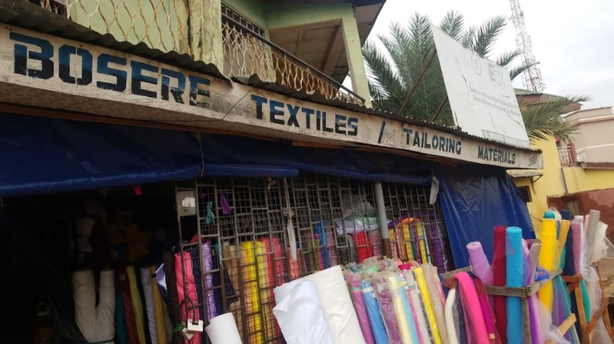 Shop signage reading Bosere Textiles / Tailoring Materials above an open shopfront stacked with rolls of fabric.